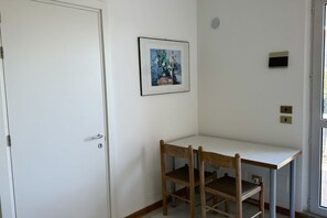 A small room with a white door, a framed picture on the wall, a white table with a chair, and a window.