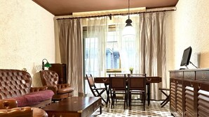 Una habitación con un sofá de cuero marrón, una mesa de centro de madera, una mesa de comedor con sillas, un televisor sobre un mueble bajo y una ventana con cortinas.