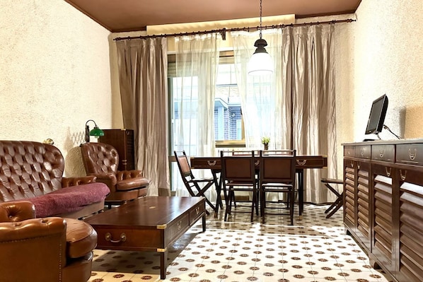 A room with a brown leather sofa, a wooden coffee table, a dining table with chairs, a TV on a stand, and a window with curtains.