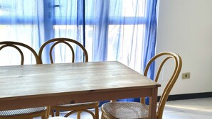 A dining table with four chairs, a window with blue curtains, and a light switch on the wall. - Palme 10 - Marcelli, two-room apartment with garden, Residence Le Palme (Marcelli)