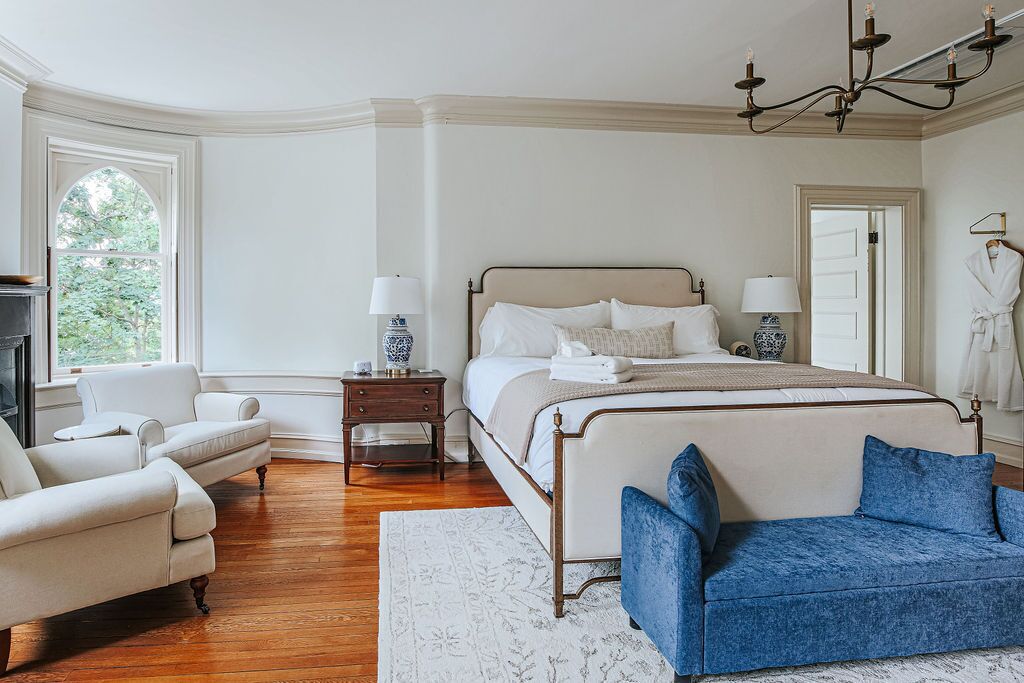 A bedroom with a large bed, two bedside tables, a chandelier, a window, and a blue armchair.