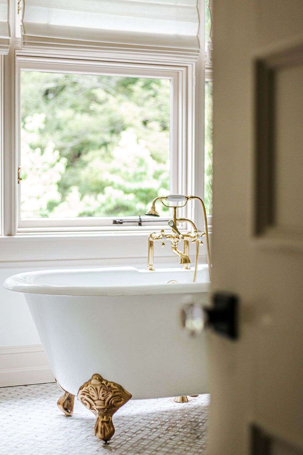 A white freestanding bathtub with gold fixtures in a bathroom with a large window.