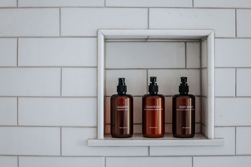 Three bottles of shampoo, conditioner, and body wash in a white shelf against a white tiled wall.