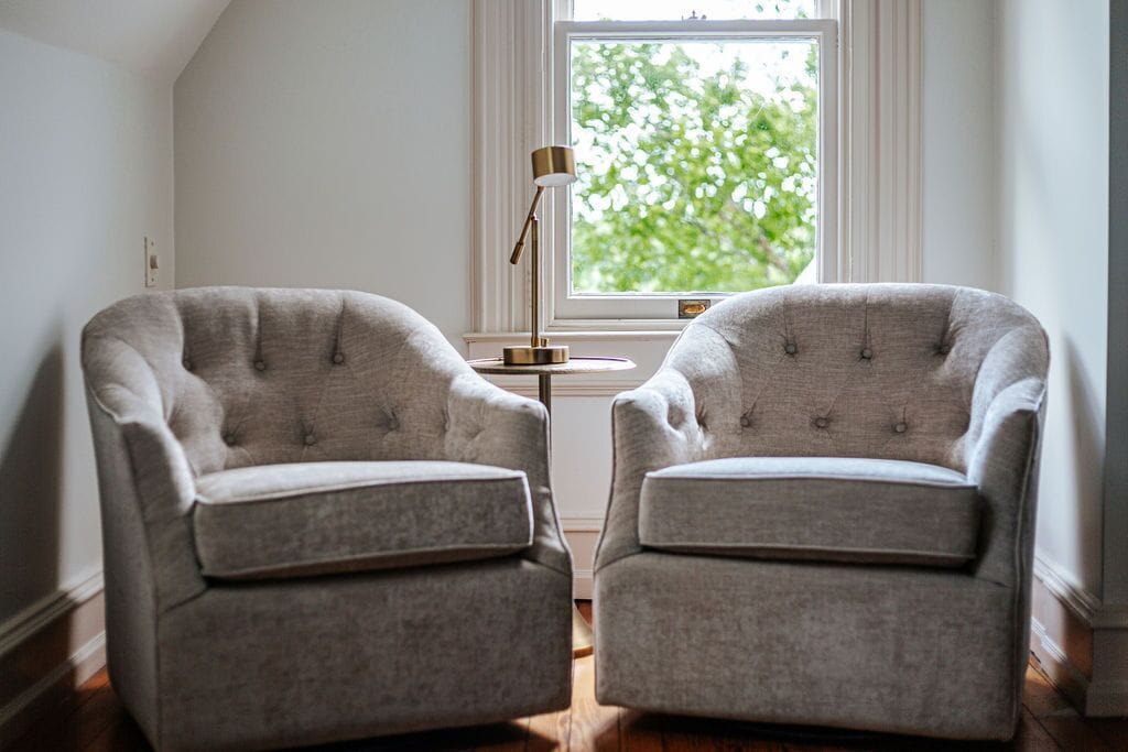 Dos sillones en una habitación con ventanal, una mesita con lámpara y vista a zonas verdes.