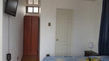 Un dormitorio con cama, televisor montado en la pared, armario, puerta y mesita de noche con lámpara.