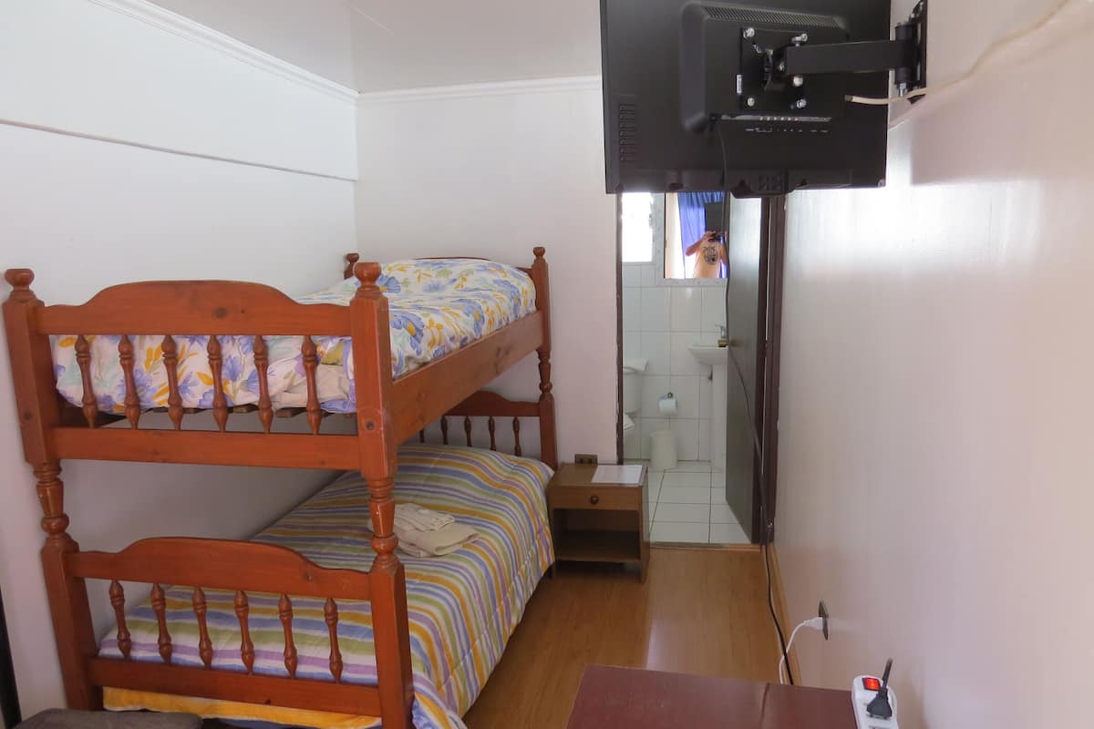 Una habitación con una cama litera, una mesita pequeña, un televisor montado en la pared y un baño visible a través de una puerta abierta.
