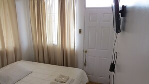 A bedroom with a bed, curtains, a door, and a wall-mounted TV. - Palm Hostal (Iquique)