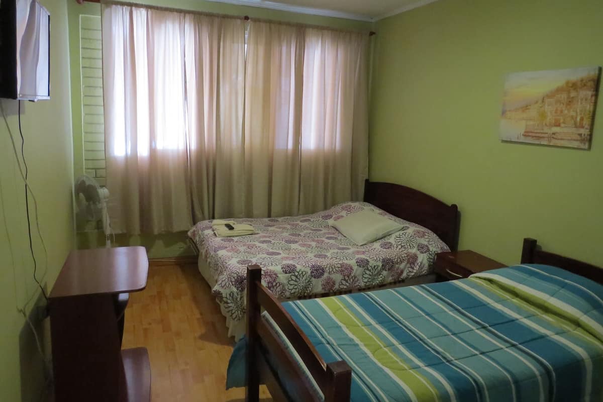 Un dormitorio con dos camas, un escritorio, una silla, una ventana con cortinas y un televisor fijado a la pared.