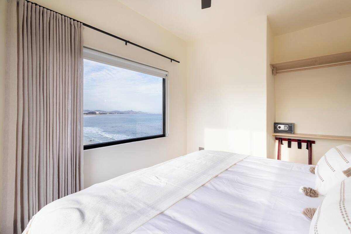 Une chambre d’hôtel avec un lit, une fenêtre avec des rideaux, une table de chevet avec une radio et une vue sur la mer.