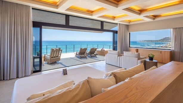 A modern hotel room with a large bed, a seating area with chairs, a wooden table, and a view of the ocean. - Cabo Break Hotel (San José del Cabo)