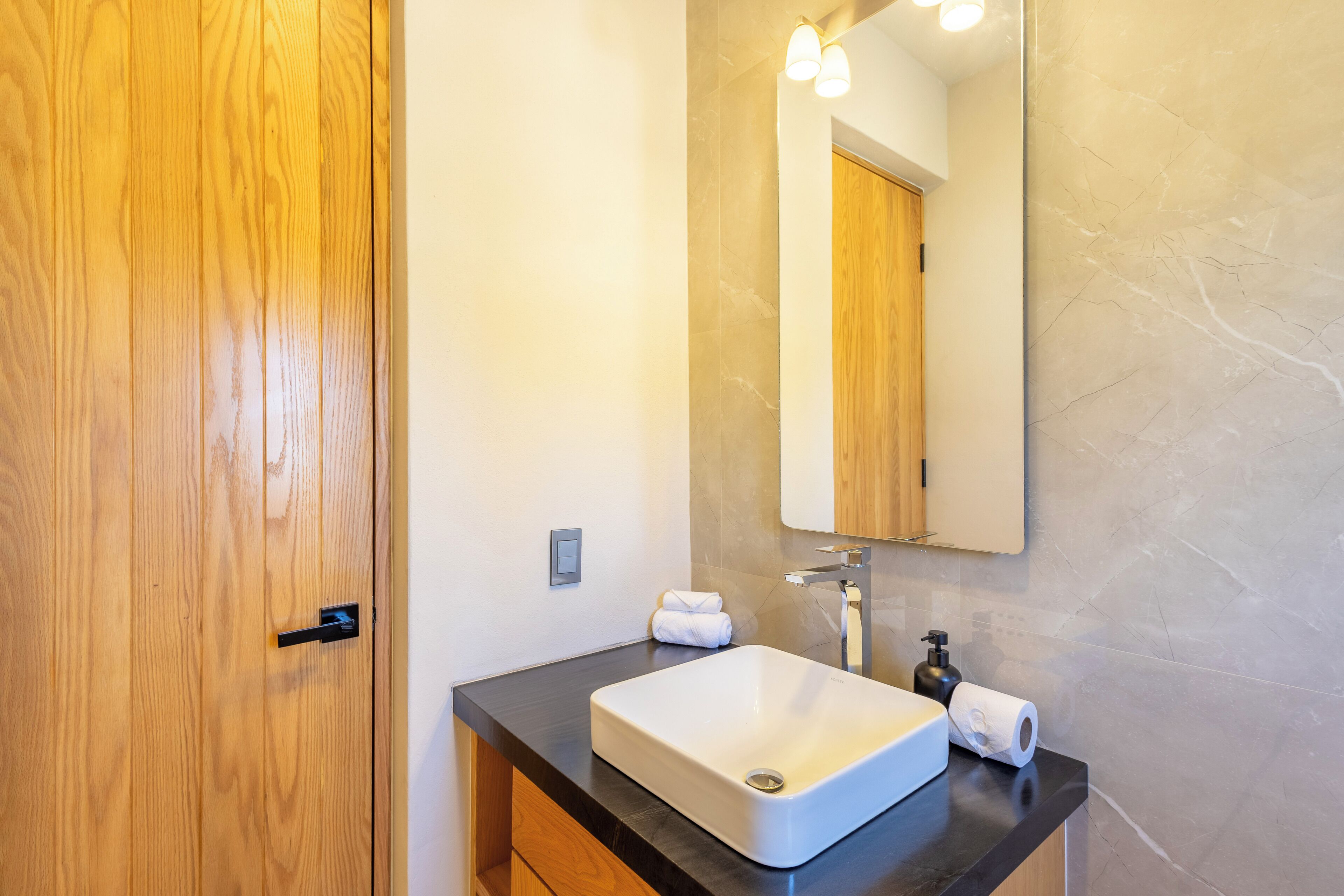 A modern bathroom with a wooden door, a black countertop, a white sink, a mirror, and a towel.