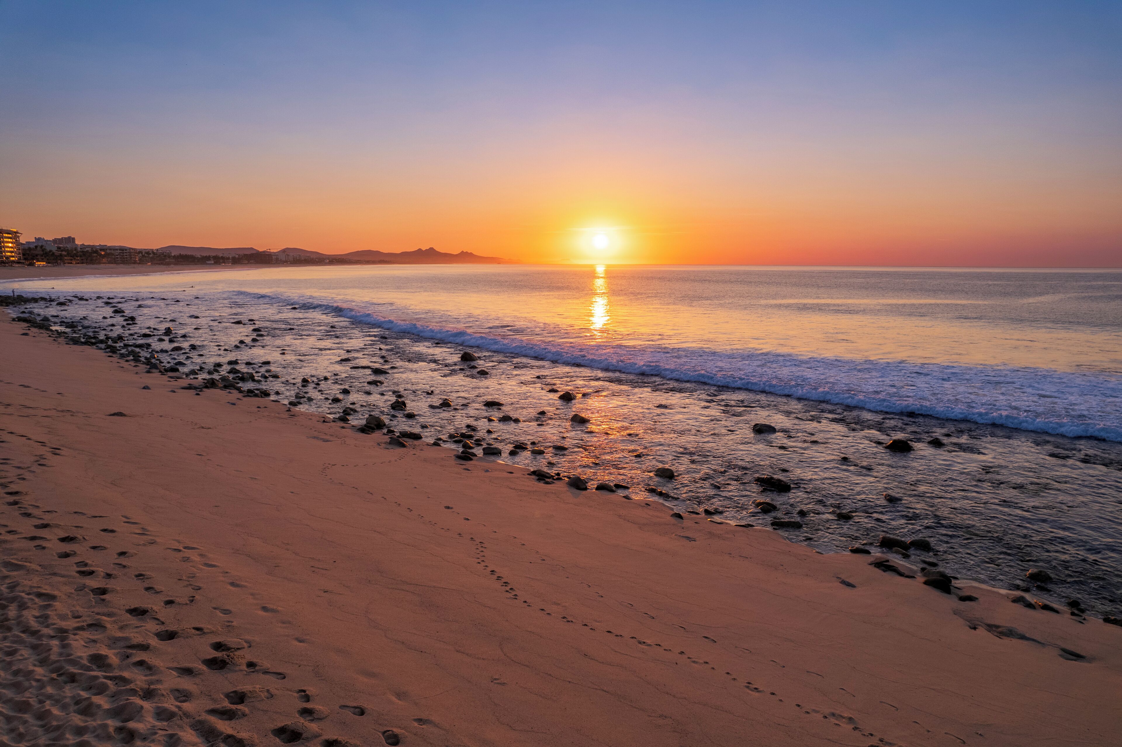 A beach with footprints leading to the water, a sunset in the background, and buildings in the distance.