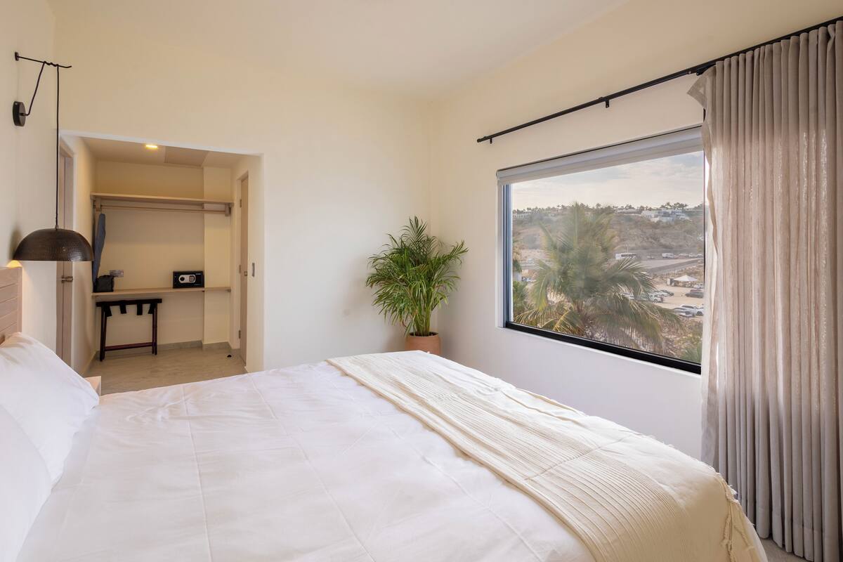 Une chambre à coucher avec un grand lit, une plante en pot, un bureau et une vue sur un paysage par la fenêtre.