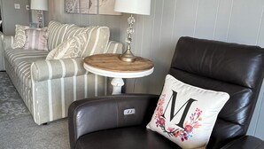 A living room with a dark leather armchair, a striped sofa, and a small round table with a lamp. There are framed pictures on the wall and a carpeted floor. - Miele di North Myrtle Beach — 1st Floor, 2BR-2BA Condo @ Inlet Point Villas (North Myrtle Beach, SC)