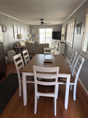 Un coin repas avec une table en bois et des chaises blanches, un canapé, un téléviseur et un ventilateur de plafond.