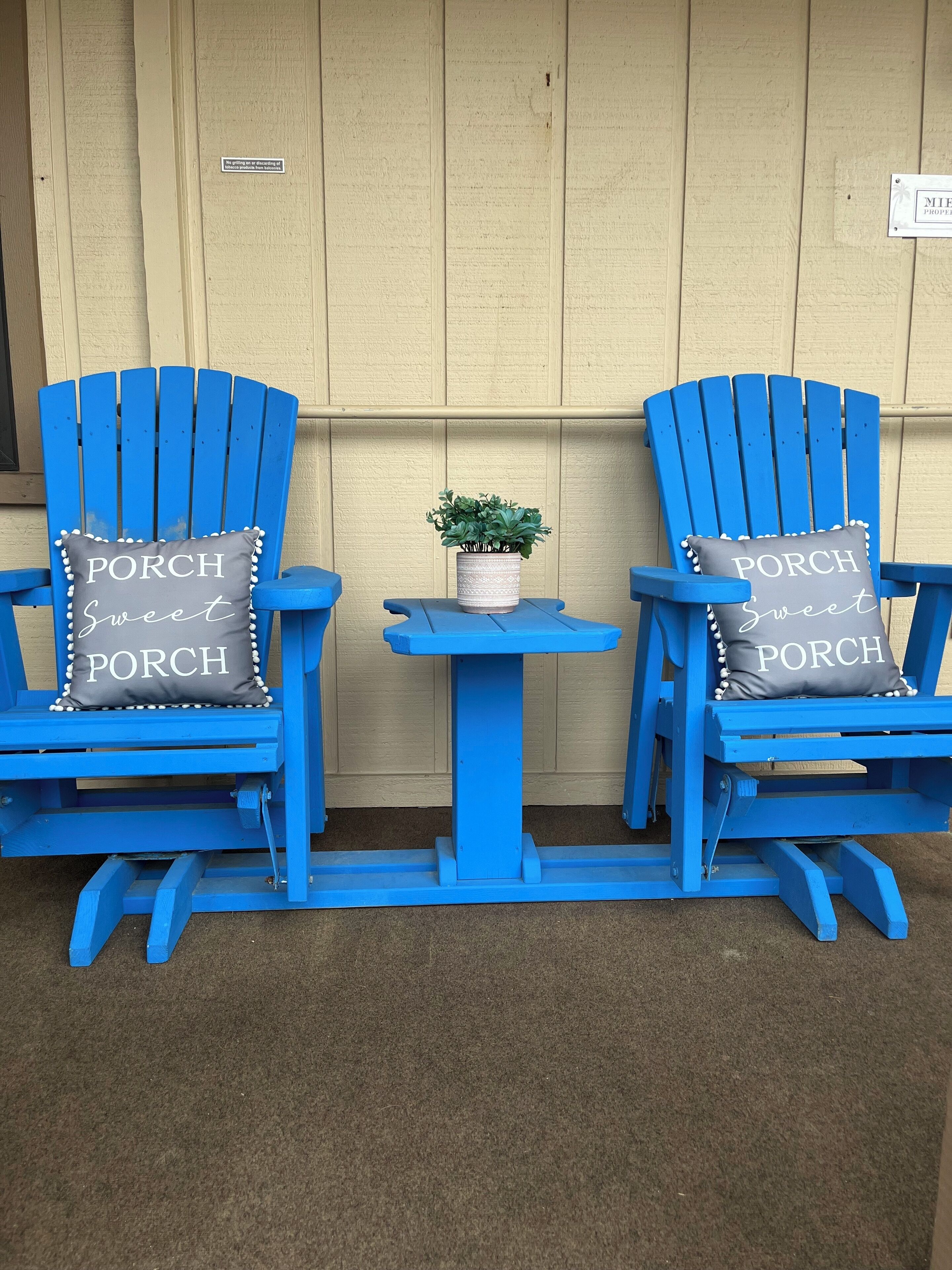 Two blue Adirondack chairs with 'PORCH Sweet' pillows, a small table with a plant, and a beige wall in the background.