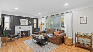 A modern living room with a fireplace, a brown leather sofa, a coffee table, and a large window with curtains. - The Drift House- Spacious Boho Family Retreat- 4 bds, 2 baths, 6 TVs, Movie Room (Clinton Township)