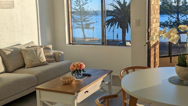 A living room with a sofa, a coffee table with a vase of flowers, and a view of a body of water through large windows. - Marine Parade - Ocean Views - Unit 6 (The Entrance)