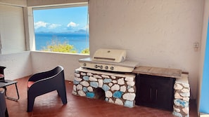 Zona de barbacoa exterior con chimenea de piedra, una silla negra y vista al mar.