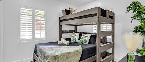 A bunk bed with a checkered rug, a bedside table with a vase, and a window with blinds.