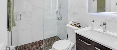 Un baño moderno con ducha de vidrio, inodoro blanco, lavabo con espejo y mueble de madera oscura.