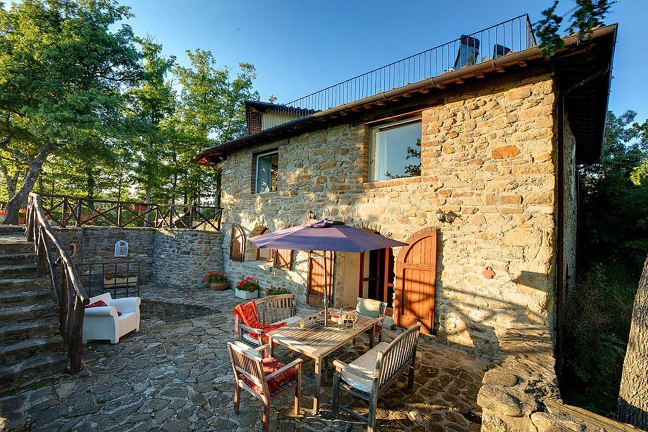 Una casa de piedra con balcón, una mesa de madera con sillas y una escalera que lleva al piso superior.