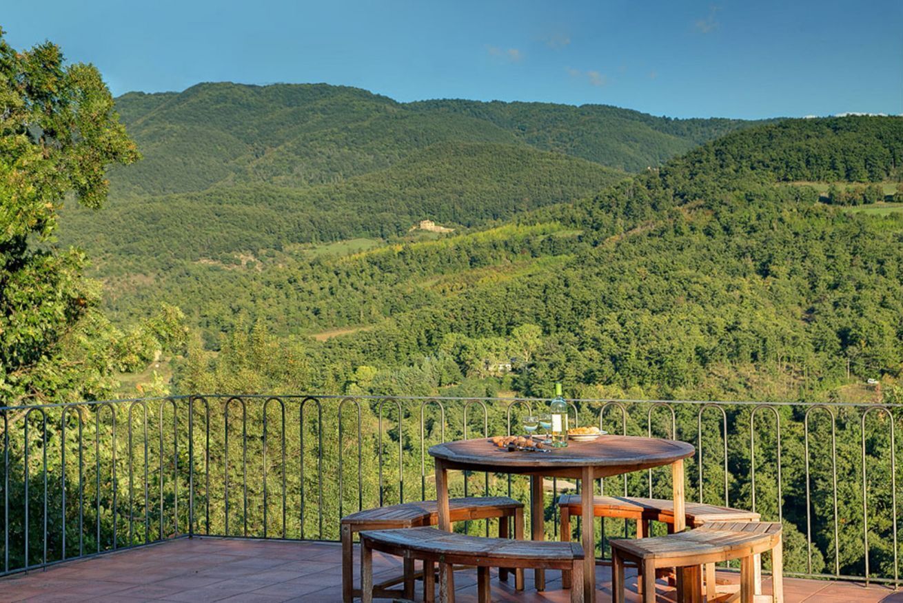 Terraço com mesa redonda de madeira e bancos, rodeado por guarda-mão de metal, com vista para um vale florestado.