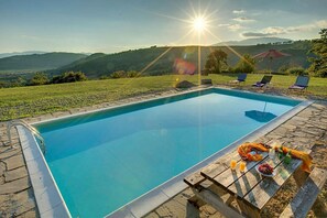 A swimming pool with a stone border, lounge chairs, and a picnic table with food and drinks.