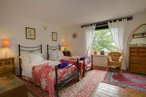 Ein Schlafzimmer mit zwei Betten, einem Fenster mit Vorhängen, einem Sessel, einer Kommode und einem Nachttisch mit Lampen.