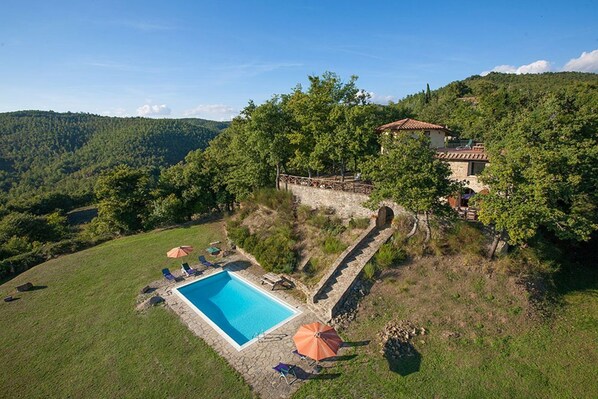 A swimming pool with lounge chairs and umbrellas, surrounded by a stone wall and trees, with a house and hills in the background.