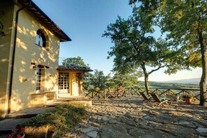 A stone patio with outdoor seating, a building with a tiled roof, and a view of the landscape.