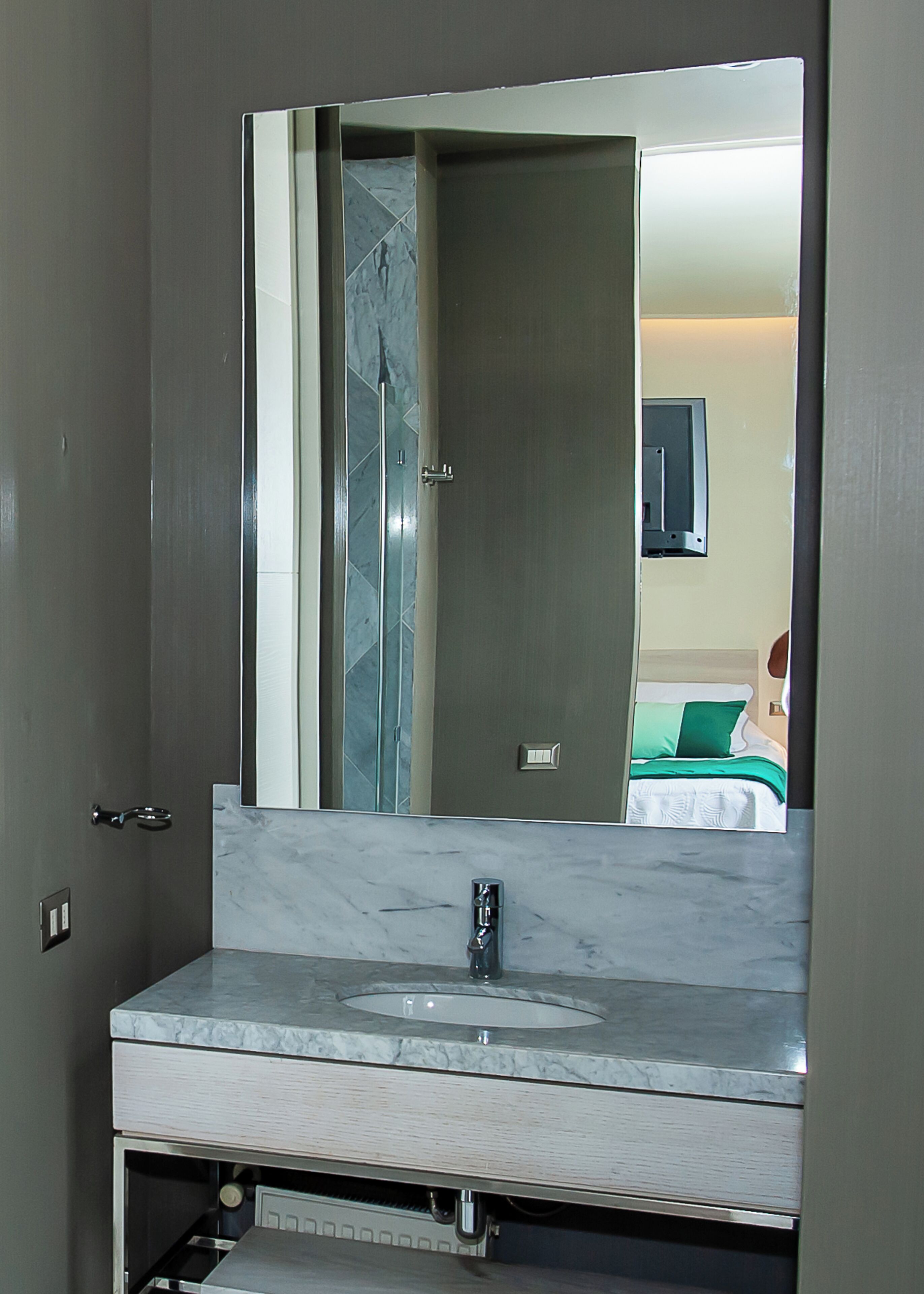 A bathroom with a marble sink, a large mirror, and a door leading to a bedroom with a bed and a green pillow.