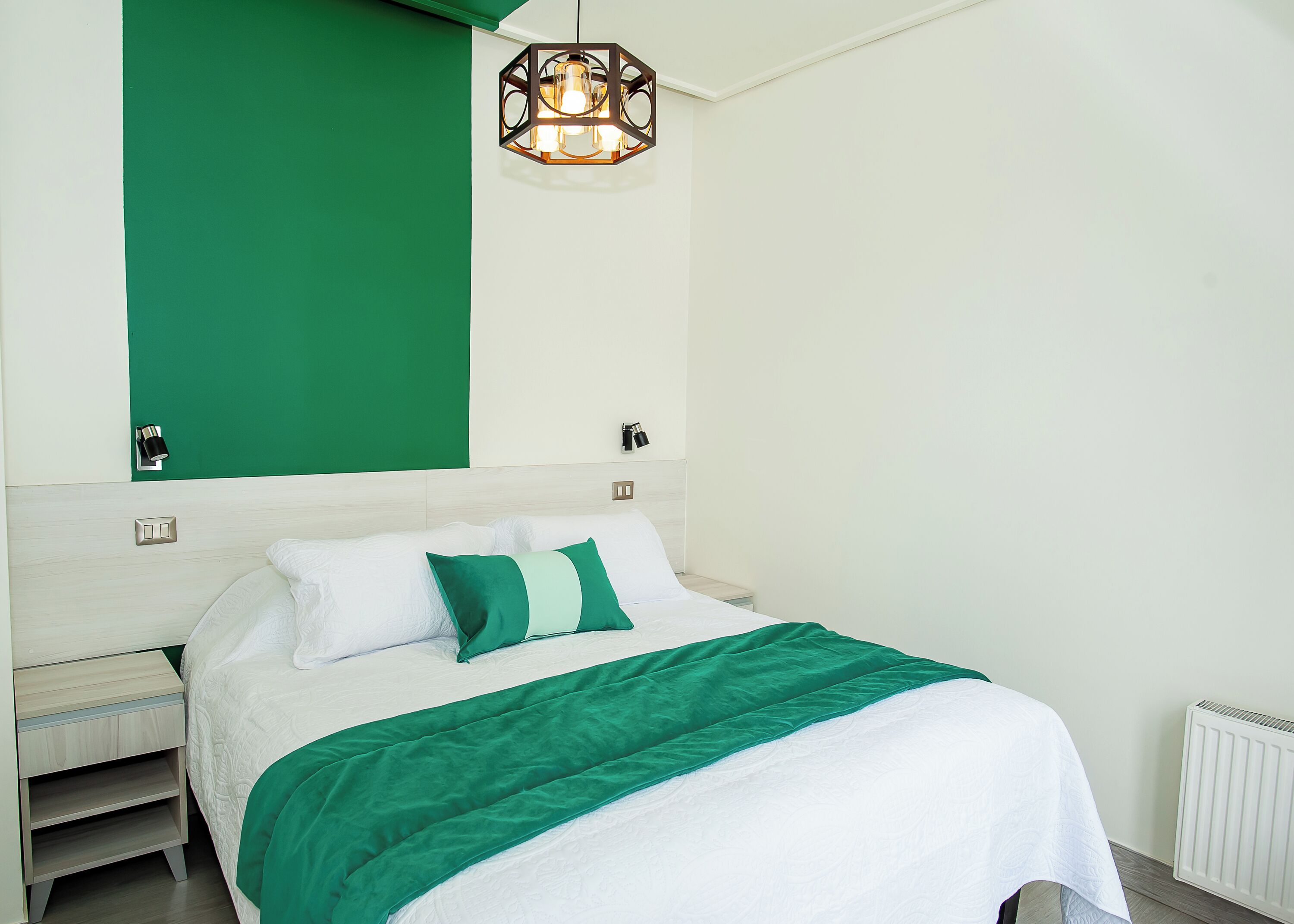 Un lit bien fait, recouvert d’une couvre-lit verte et blanche, agrémenté d’un plaid assorti, et surmonté d’un luminaire suspendu au design moderne.