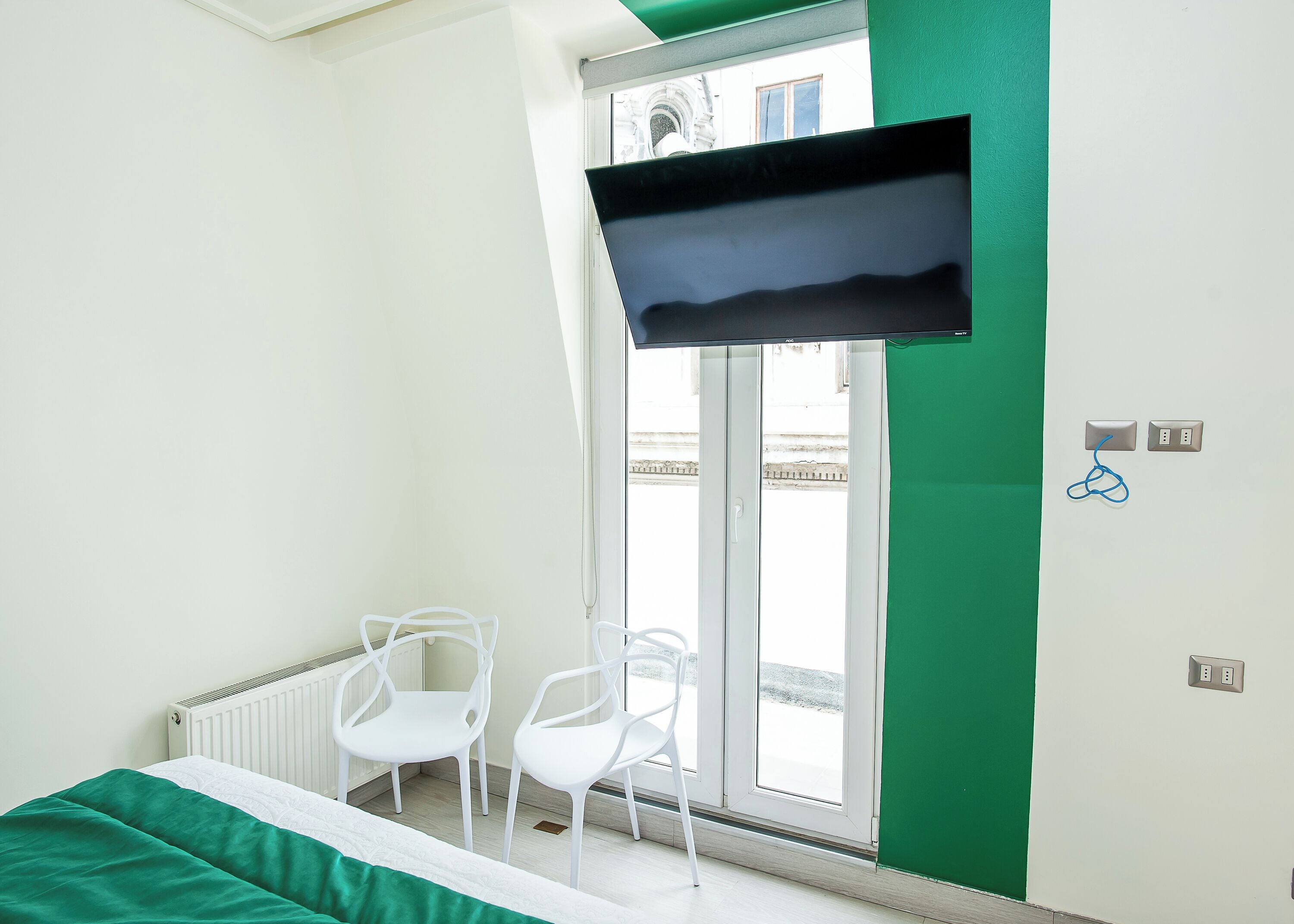 A hotel room with a bed, two white chairs, a TV mounted on the wall, a window with a view of a building, and a green accent wall.