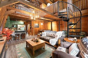A rustic cabin interior with a dining area, a living room with a sofa and coffee table, and a spiral staircase.