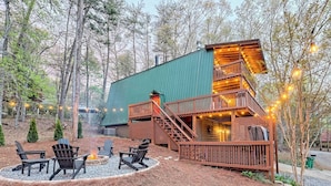 A cabin with a green roof, wooden deck, and outdoor seating area with chairs and a fire pit.