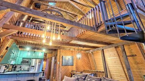 A rustic cabin interior with a kitchen, dining area, and a loft bedroom with a balcony.