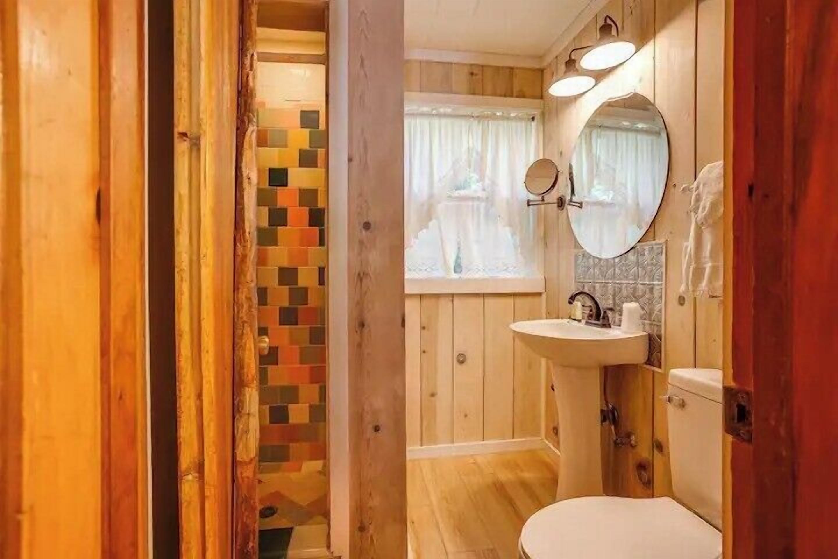A wooden interior with a wooden floor, a white toilet, a sink with a mirror, and a window with curtains.