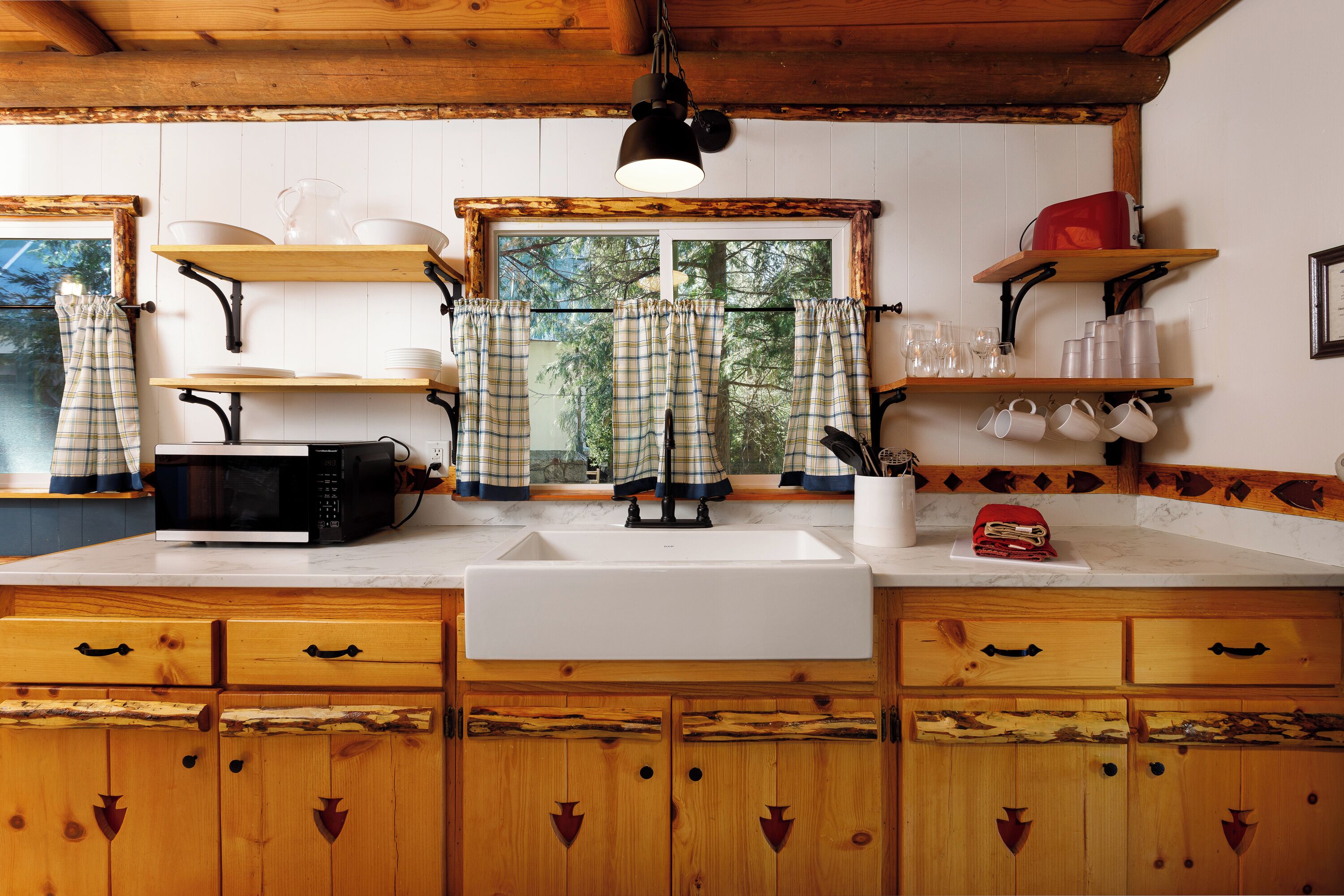 A kitchen with wooden cabinets, a white sink, and a microwave. There are shelves with dishes and a window with curtains.