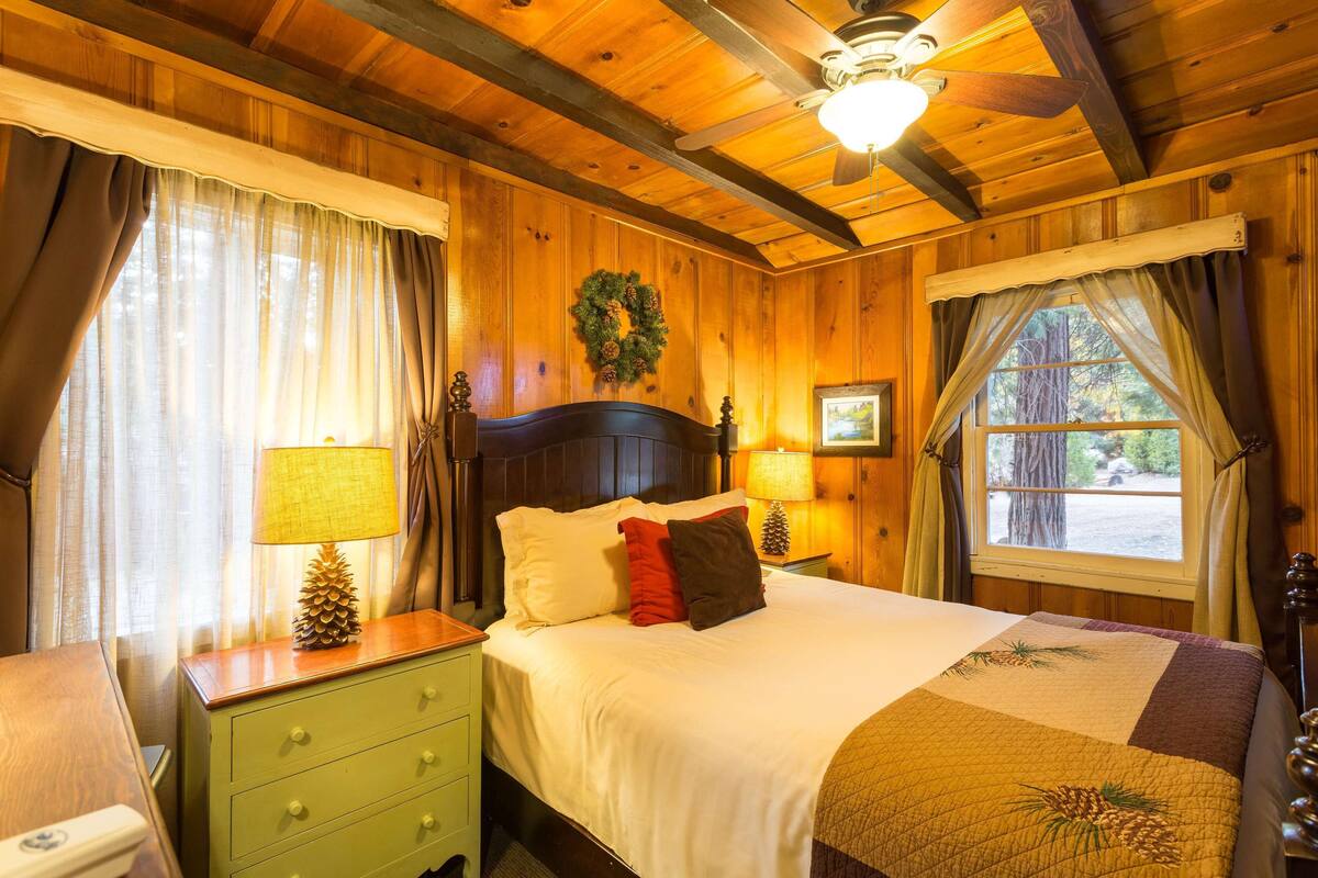 Une pièce chaleureuse, avec des murs en bois, comprenant un lit, une table de chevet, une lampe et une fenêtre avec des rideaux.