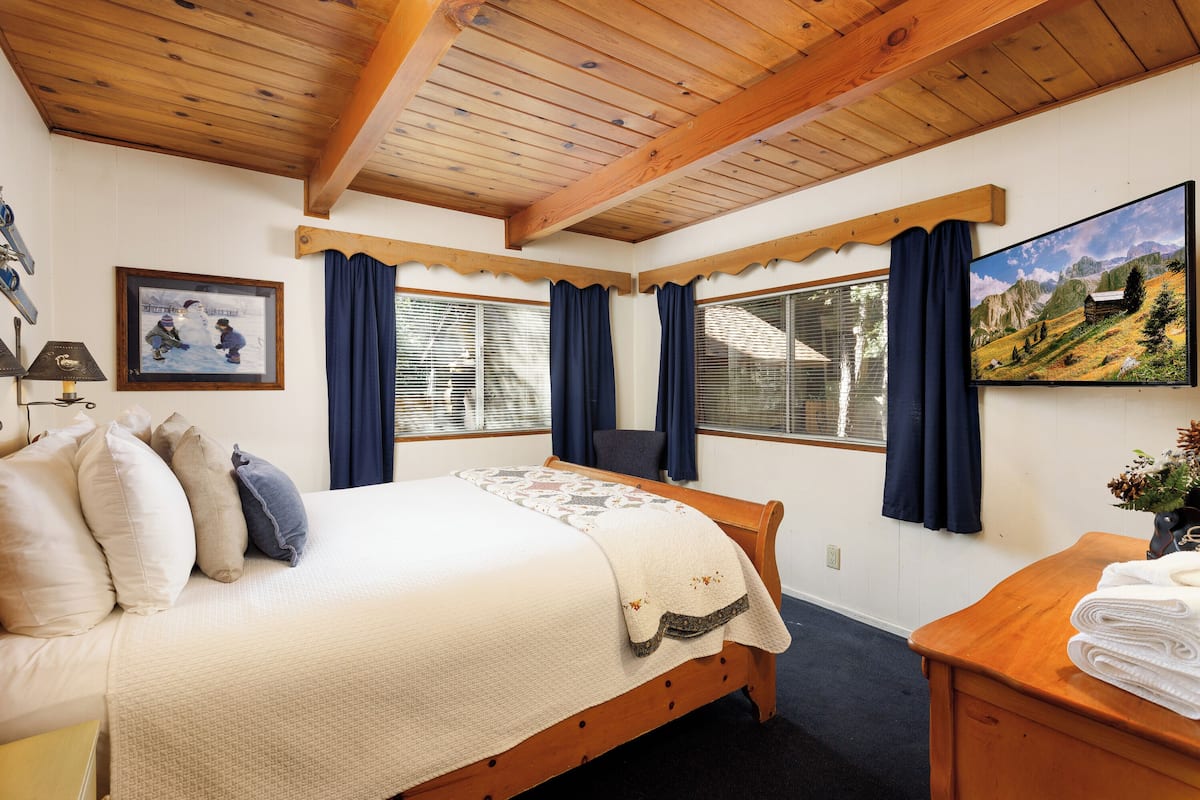 Une chambre à coucher avec un lit en bois, un tableau encadré, un bureau avec une lampe et une fenêtre avec des rideaux.