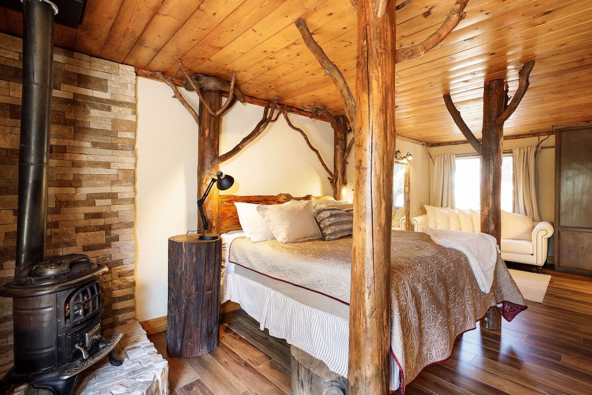 Une chambre à coucher rustique avec un plafond en bois, une cheminée en pierre, un lit recouvert d’une courtepointe blanche et marron, et un fauteuil blanc.
