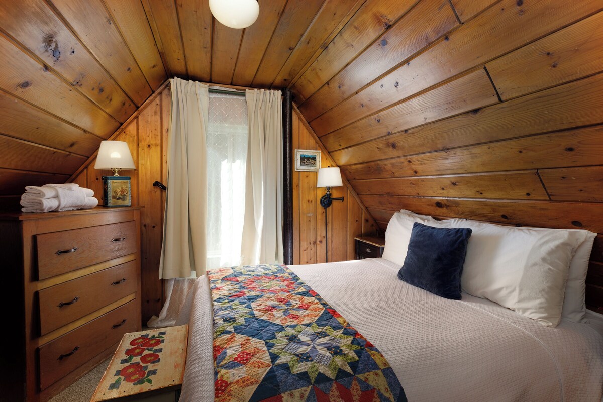 A cozy attic bedroom with wooden walls, a bed with a quilt, a dresser, and two wall lamps.