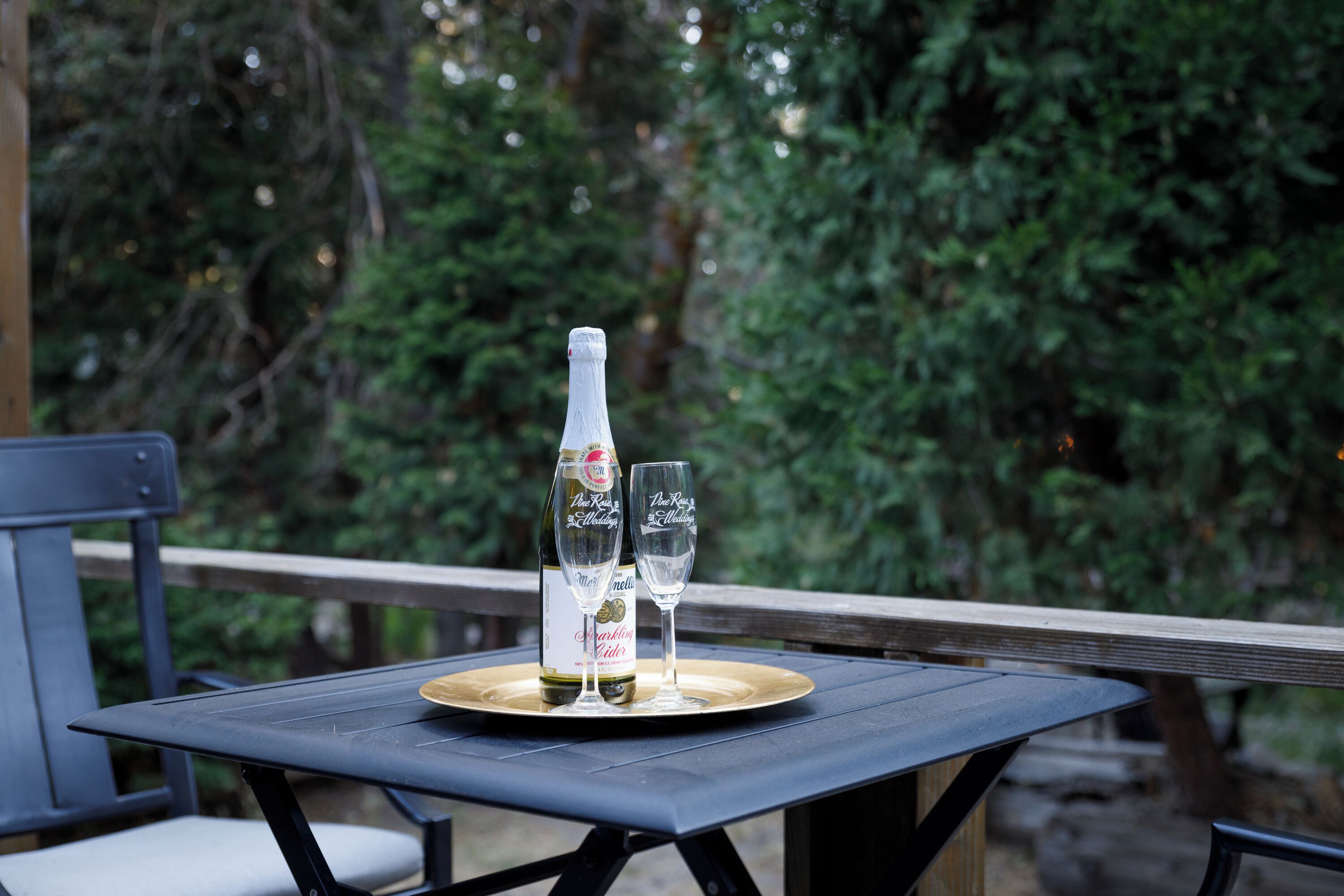 A table with a bottle and two glasses on a tray, set on an outdoor deck with a wooden railing and greenery in the background.