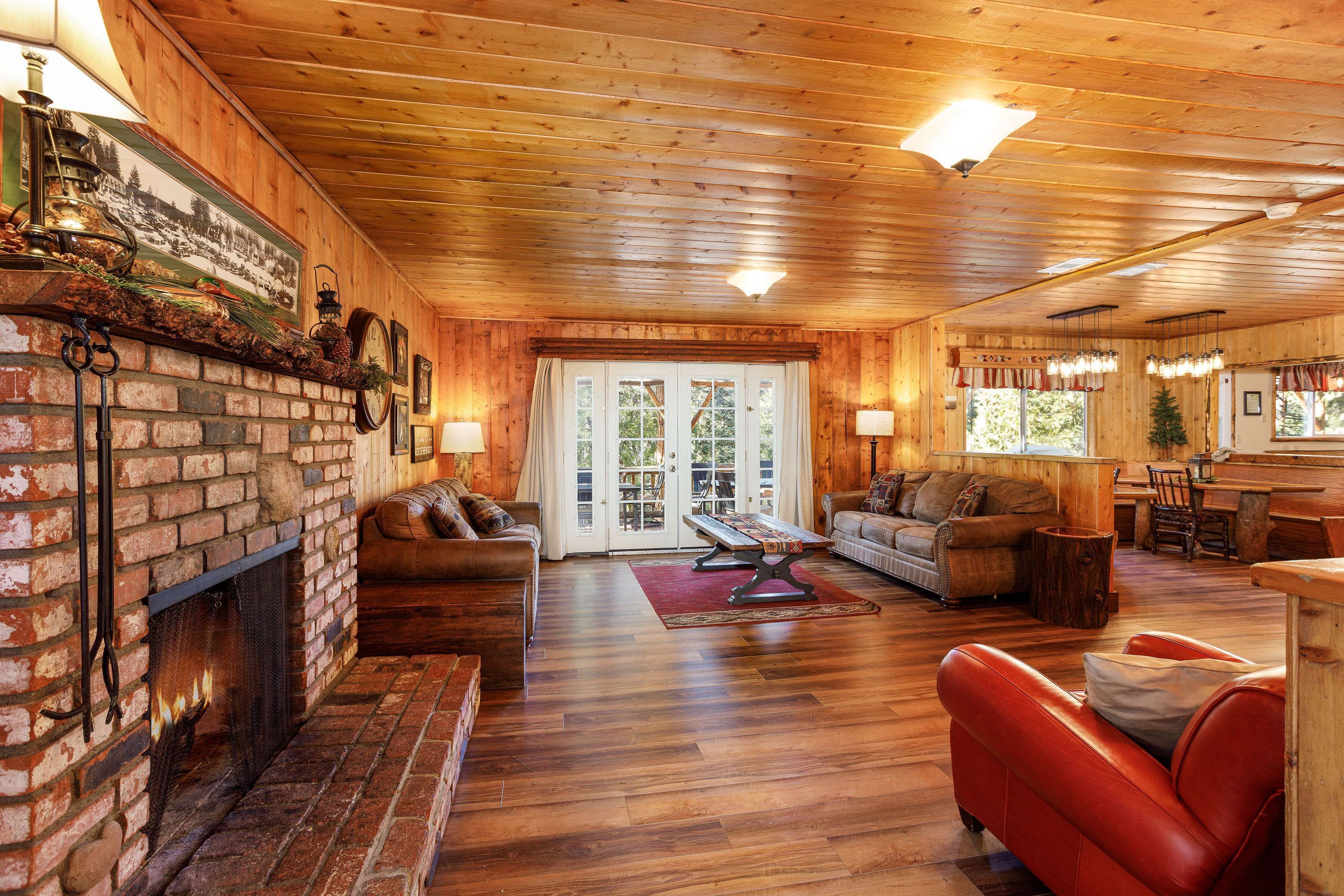 A cozy cabin interior with a fireplace, wooden floors, a dining area, and a living room furnished with leather sofas and a red armchair.