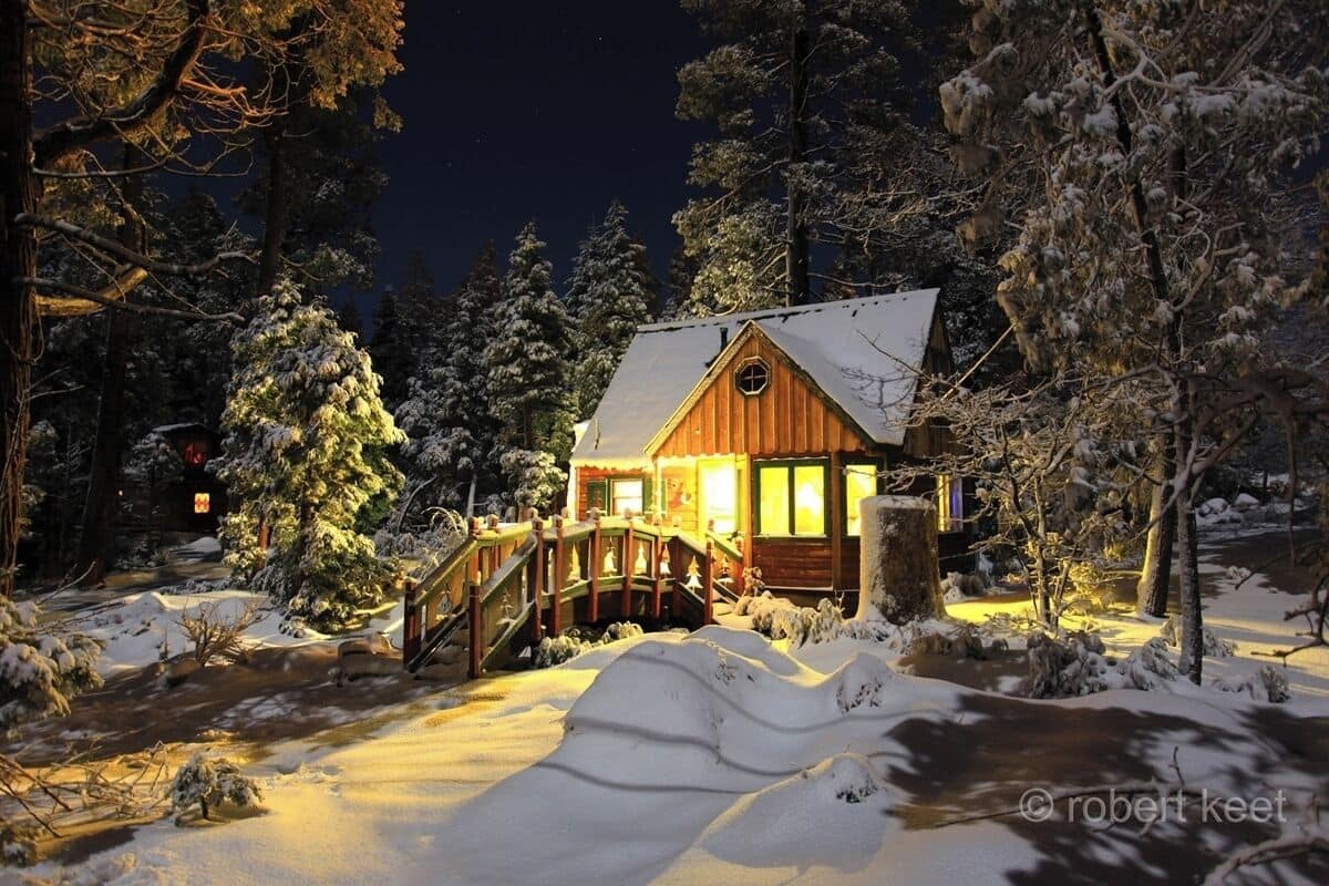 Une cabane aux murs en bois, un pont couvert et des arbres recouverts de neige.
