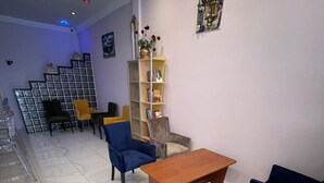Amplia sala de estar con piso de baldosas, una mesa de centro de madera y varias sillas. Se observa una estantería con libros y objetos decorativos, además de un cuadro en la pared.
