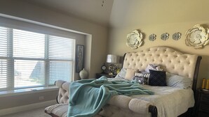 A bedroom with a large bed, a bench, a window with blinds, and decorative wall art. - Charming Crowley Rental (Crowley)