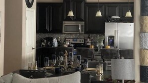A modern kitchen with dark cabinetry, a microwave, and a refrigerator. There are pendant lights above the counter and a dining area with a table set for two. - Charming Crowley Rental (Crowley)