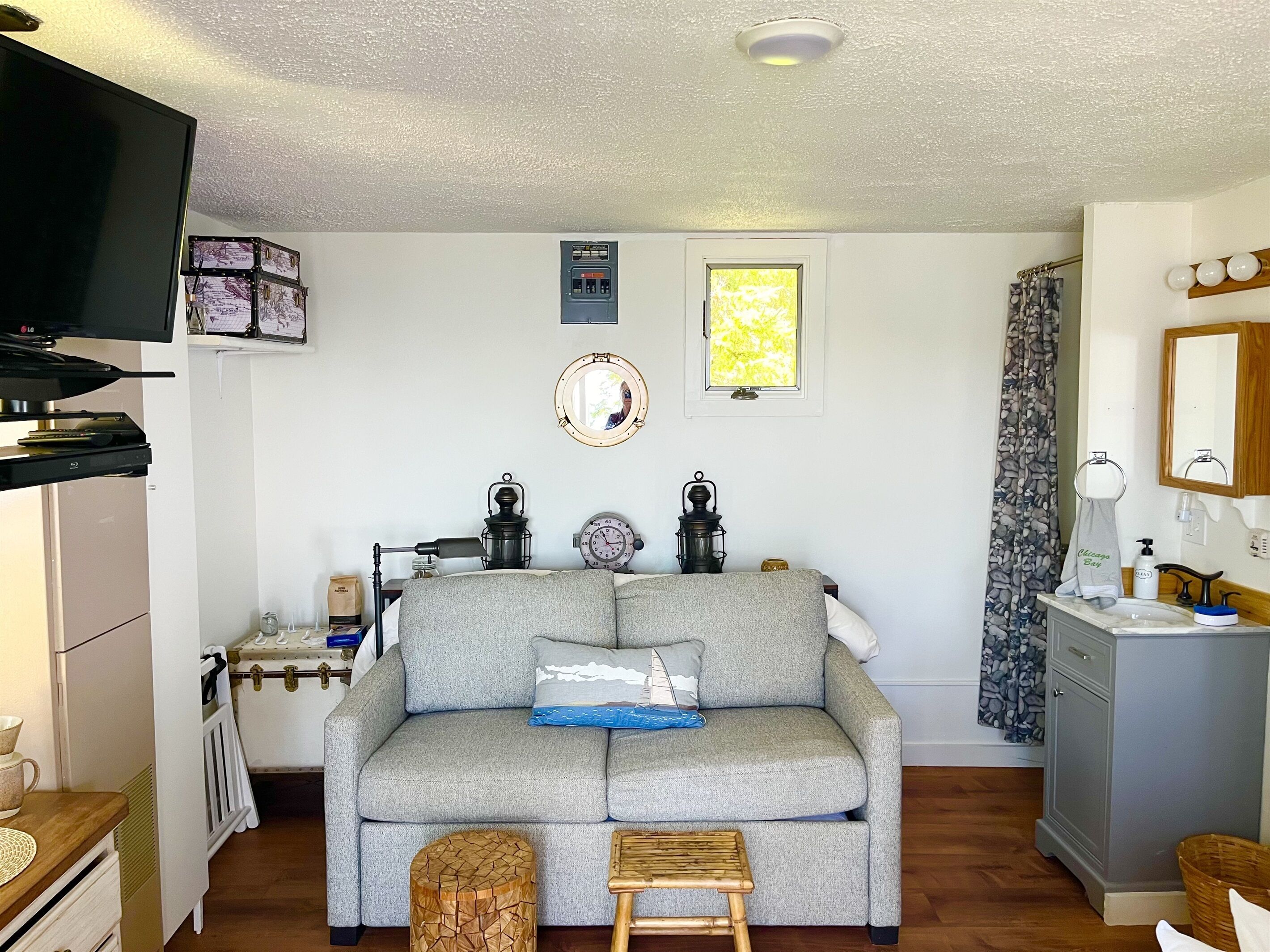 A cozy living room with a grey sofa, a small wooden stool, a coffee table, a television, a fireplace, and a window with a view of trees.